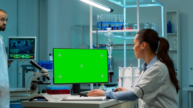 Foto científico sentado en el escritorio trabajando en una computadora personal con pantalla verde de maqueta. en segundo plano, el investigador de laboratorio del hombre discutiendo con el médico sobre el desarrollo de la vacuna trayendo muestras de sangre