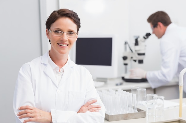 Científico sonriendo a la cámara brazos cruzados y otro trabajando con microscopio