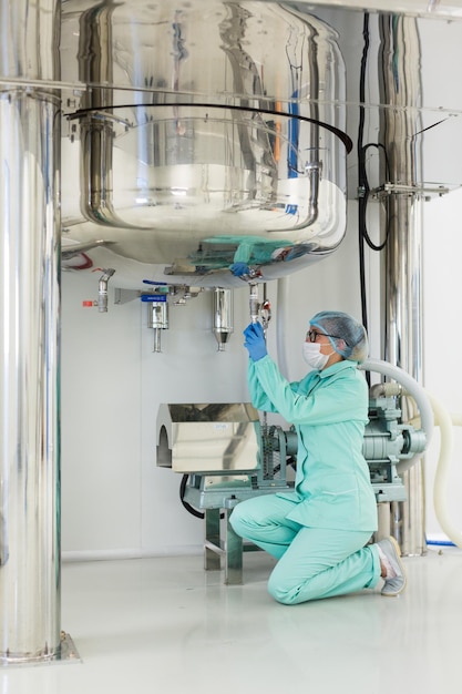 El científico con traje de laboratorio azul se sienta bajo la presión de control de un gran barril de acero y se sienta de rodillas