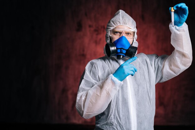 El científico con traje protector, gafas y respirador sostiene el tubo de ensayo en la mano y lo señala con el dedo. Concepto de trabajo en la vacuna contra el coronavirus. Foto de estudio sobre fondo rojo oscuro.