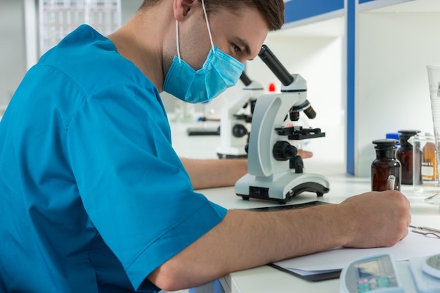 Científico en uniforme está escribiendo notas de su investigación después de mirar a través de un microscopio en un laboratorio. Concepto de salud y biotecnología