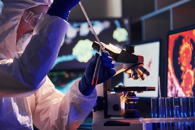 Científico con uniforme protector blanco cerca de pantallas con datos trabaja con coronavirus y tubos de sangre en laboratorio