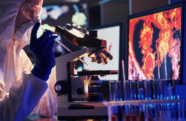 Científico con uniforme protector blanco cerca de pantallas con datos trabaja con coronavirus y tubos de sangre en laboratorio