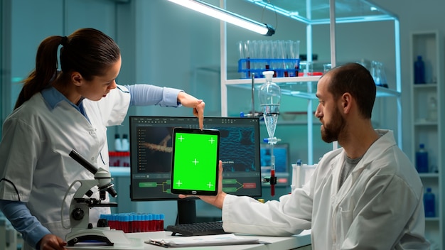 Los científicos de investigación médica que utilizan el bloc de notas con pantalla verde simulan la plantilla en el laboratorio de ciencias aplicadas apuntando en la pantalla de croma. Los ingenieros de laboratorio con batas blancas realizan experimentos trabajando