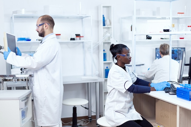 Cientista africano trabalhando no computador usando equipamento de proteção, fazendo análise. Equipe multiétnica de pesquisadores trabalhando em solução de testes de laboratório de microbiologia para fins médicos.