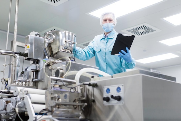 Cientista caucasiano em uniforme de laboratório azul verifica leituras de grande máquina cromada de aço com eixos olhe para a câmera