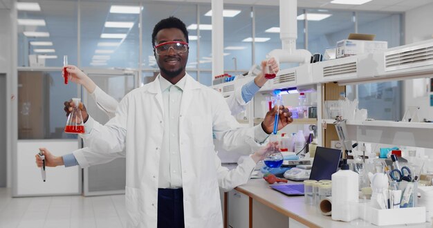 Cientista de laboratório afro com várias mãos em jaleco e óculos de segurança segurando frascos e tubos de ensaio