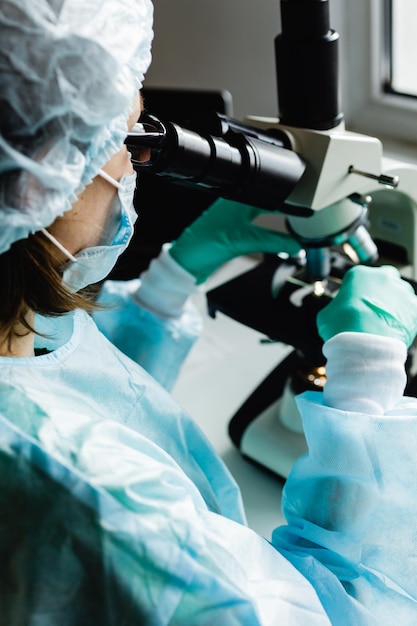 Cientista em luvas médicas azuis e amostras de aprendizagem uniformes com microscópio em laboratório