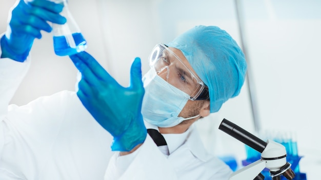 cientista em roupas protetoras, olhando para o líquido no frasco.