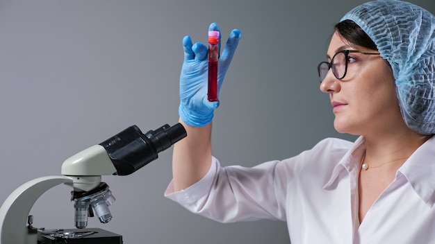 Cientista experiente com óculos olha para uma amostra de líquido vermelho em um tubo de ensaio perto do microscópio, trabalhando em um hospital moderno.