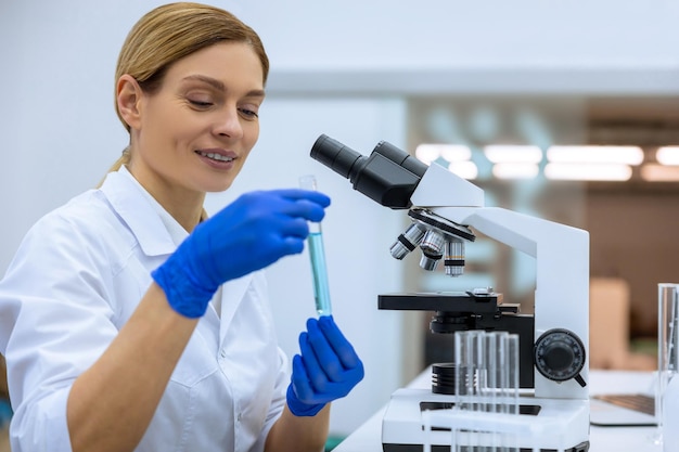 Cientista feminina atraente e positiva examinando líquido em tubo de ensaio em laboratório fazendo pesquisa de DNA posando em seu local de trabalho