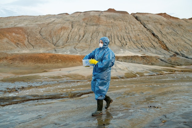 Cientista feminina contemporânea usando óculos de proteção, respirador, luvas de borracha e botas carregando um recipiente de plástico nas colinas e no céu