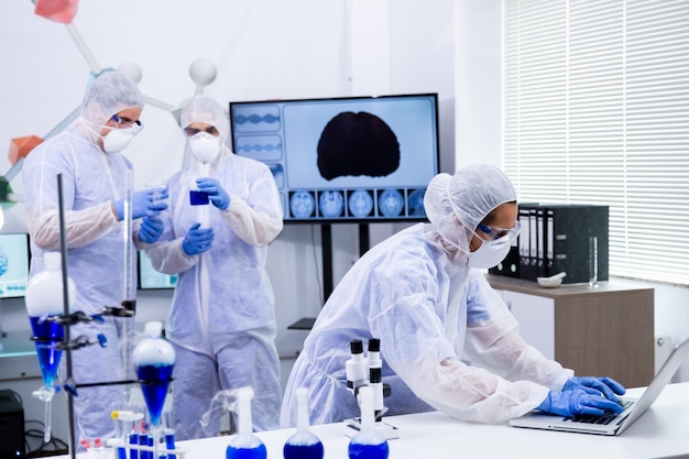 Foto cientista feminina trabalhando em laptop e colegas trabalhando com solução azul reativa em um laboratório de pesquisa moderno.