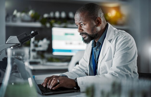 Cientista homem laptop e fazendo pesquisas em um laboratório com tecnologia da informação e análise de dados Pessoa negra ou médico digitando online à noite para desenvolvimento de ciências médicas na internet