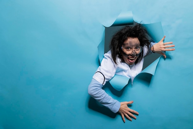 Cientista humorístico tolo com expressão cômica sobre fundo azul. Jovem químico adulto com olhar maluco, cabelo bagunçado e cara suja agindo pateta após a explosão do experimento. Tiro de estúdio.