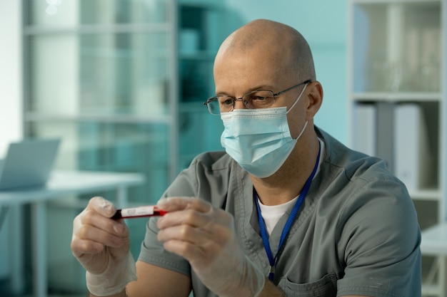 Cientista maduro e careca usando máscara protetora e luvas, olhando para uma amostra de sangue em um frasco pequeno durante uma pesquisa científica em um laboratório químico