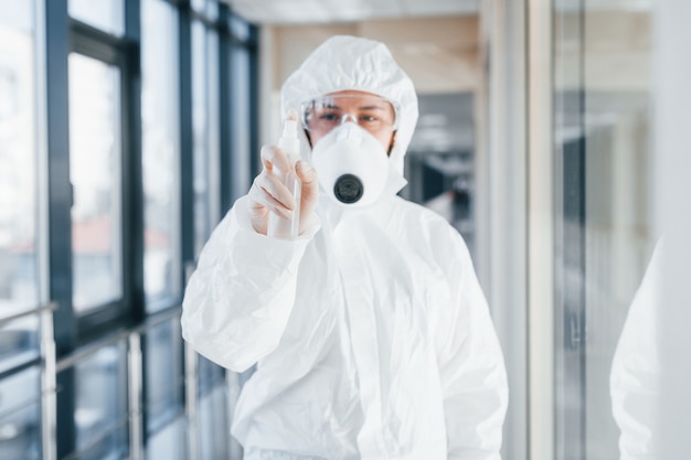 Cientista médica no jaleco, óculos de proteção e máscara em pé dentro de casa com spray antibacteriano