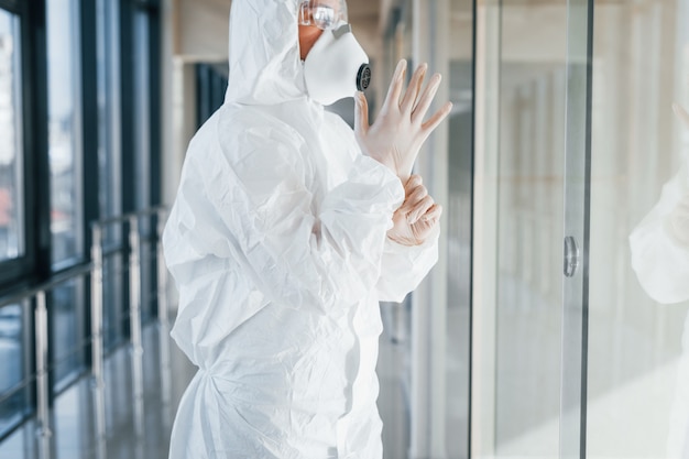 Cientista médica no jaleco, óculos defensivos e máscara de pé dentro de casa e usando luvas