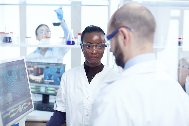 Cientista médico africano sério discutindo sobre o experimento de vírus com colegas sentados em frente ao monitor do computador. Equipe multiétnica de pesquisadores médicos trabalhando juntos em uso de laboratório estéril em