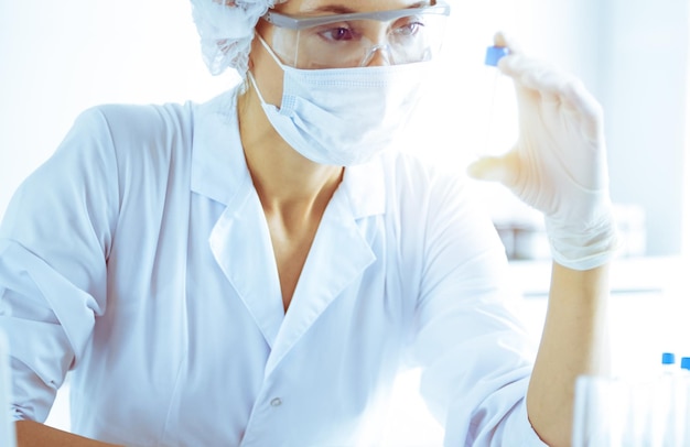 Cientista profissional feminina em óculos de proteção, pesquisando tubo com reagentes em laboratório ensolarado em tons de azul. Pesquisa em medicina e ciência.