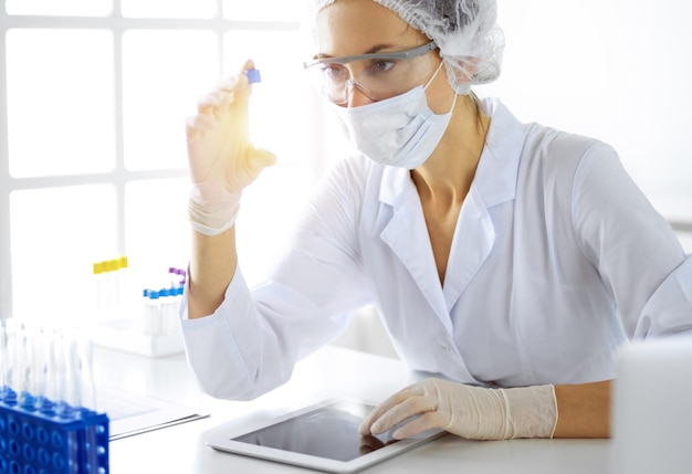 Cientista profissional feminina em óculos de proteção, pesquisando tubo com reagentes em laboratório ensolarado. Medicina e pesquisa.