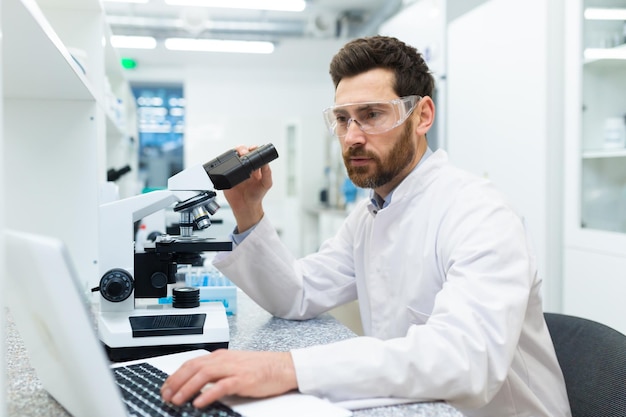 Cientista sério e atencioso trabalhando em laboratório com microscópio e laptop maduro experiente