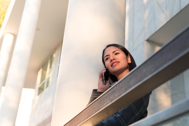 Cierra la fotografía de una joven trabajadora hablando por teléfono y usa una chaqueta gris cerca del edificio de oficinas