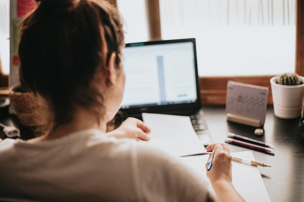 Cierra la imagen detallada de una mujer joven que estudia y trabaja en el escritorio de su casa haciendo los deberes durante la preparación universitaria para el examen con un libro de texto y tomando notas con un enfoque selectivo en el lápiz