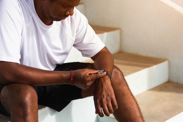 Cierra el joven atleta asiático, el corredor deportivo, el hombre negro, usa un reloj inteligente de tiempo moderno, él está sentado descansando antes de entrenar corriendo en el parque de salud de la calle al aire libre, ejercicio saludable antes del concepto de entrenamiento