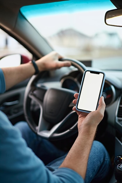 Cierra las manos de un conductor hombre irreconocible usando un teléfono móvil de maqueta de pantalla blanca mientras conduces