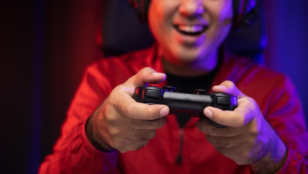 Cierra las manos masculinas sosteniendo la consola de juegos con joystick. Joven jugando videojuegos en línea. Hombre indio con chaqueta roja juega a la computadora en casa en el cuarto oscuro