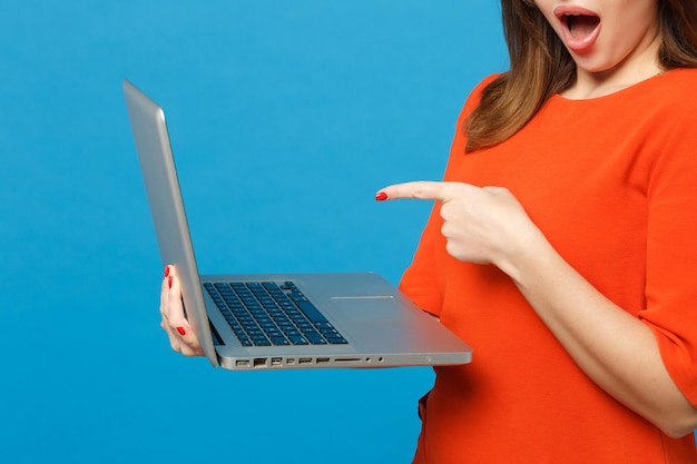 Cierra a una mujer recortada con un vestido rojo anaranjado sostenido en las manos señalando con el dedo en el ordenador portátil aislado en el fondo azul, retrato de estudio. Concepto de moda de estilo de vida de la gente. Simulacros de espacio de copia.