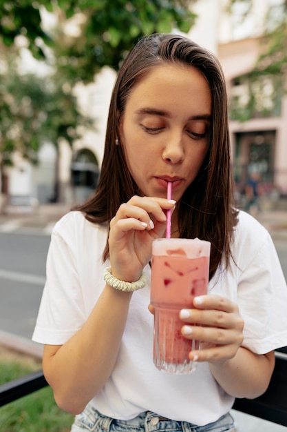 Foto cierra el retrato al aire libre de una toalla de euoepan con cabello oscuro bebiendo batido rosa de verano y descansando en la terraza de verano en la ciudad