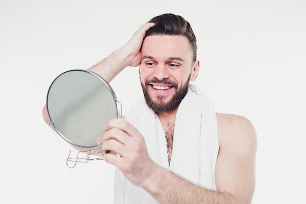 Cierra el retrato de un apuesto hombre barbudo con una toalla mirando su reflejo en el espejo del baño aislado de fondo blanco