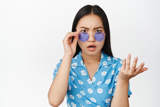 Cierra el retrato de una chica asiática con gafas de sol que se ve confundida y decepcionada, no puedo entender algo de pie sobre fondo blanco.