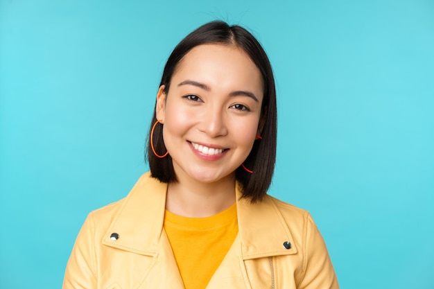 Cierra el retrato de una hermosa mujer asiática sonriente con dientes blancos mirando feliz a la cámara posando con una chaqueta amarilla sobre el fondo azul del estudio