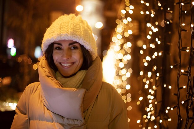 Cierra el retrato de una hermosa mujer sonriente con un sombrero blanco de invierno y una chaqueta mientras camina al aire libre con la iluminación navideña de la ciudad en el fondo y mira a la cámara