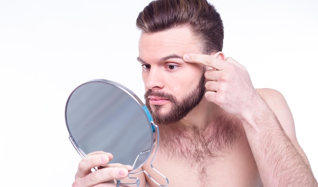 Cierra el retrato de un hombre barbudo desnudo frustrado mirándose en el espejo en busca de acné en la cara aislado de fondo blanco