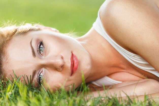 Cierra el retrato de una joven atractiva tumbada en la hierba verde en un parque durante el verano y escuchando música con sus auriculares pensativos al aire libre
