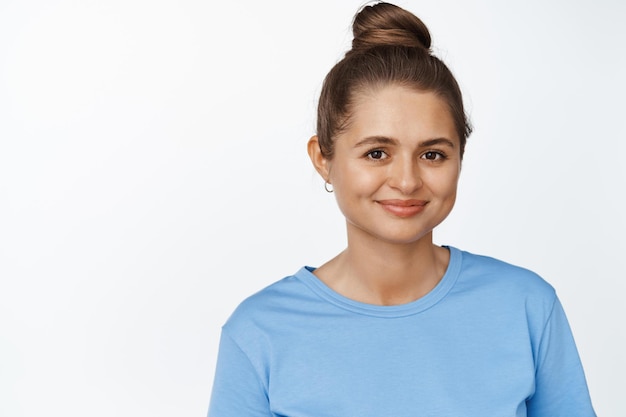 Cierra el retrato de una joven caucásica con el pelo peinado en un moño, sonriendo feliz a la cámara, de pie con una camiseta azul sobre fondo blanco.