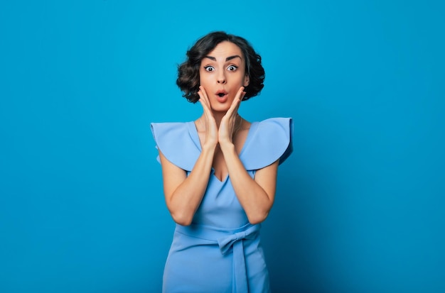 Cierra el retrato de una linda mujer sorprendida y conmocionada con un vestido azul de moda mientras sostiene sus mejillas y mira a la cámara y aislada en un fondo azul