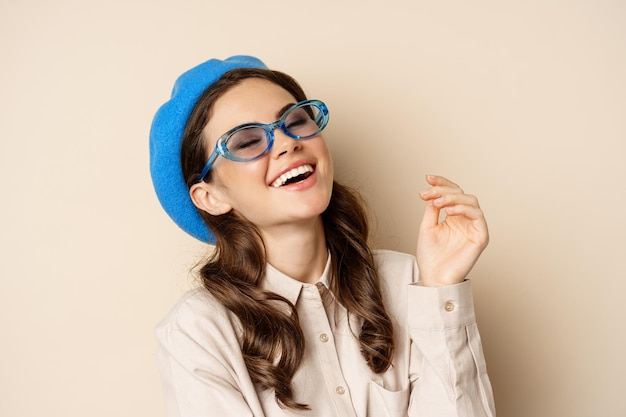 Cierra el retrato de una mujer elegante posando con gafas de sol, riendo y sonriendo, vestida con ropa de moda, de pie sobre un fondo beige