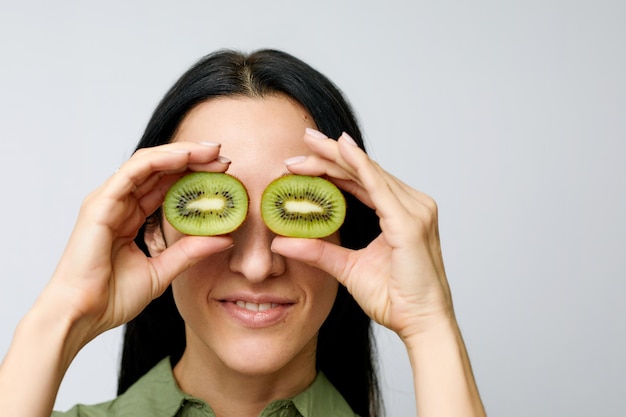 Cierra el retrato de una mujer hermosa sonriendo y sosteniendo kiwis frente a sus ojos