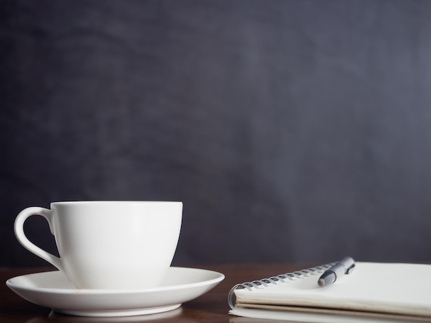 Cierra la taza de café con leche con un cuaderno y un bolígrafo sobre un fondo oscuro de la mesa de madera