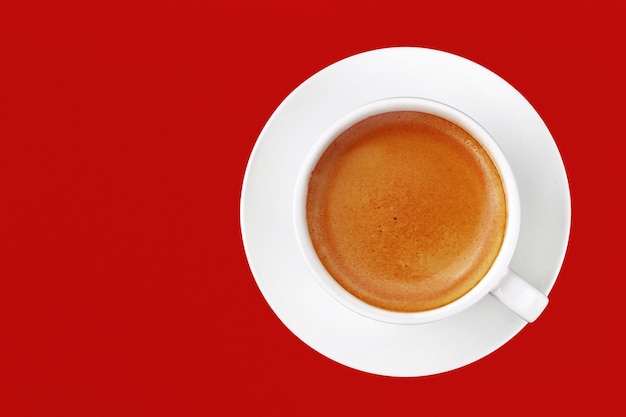 Cierra una taza llena de café espresso y un platillo blanco sobre un fondo rojo vivo, vista superior elevada, directamente arriba