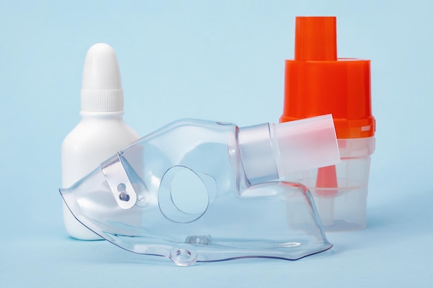 Cierre de mascarilla respiratoria, vaso atomizador y líquido en botella blanca para terapia de inhalación para asma y enfermedades respiratorias sobre fondo azul.