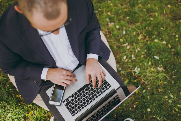 Cierre recortado de manos en el teclado. Hombre de negocios en traje clásico, gafas. El hombre se sienta en un puf suave, trabaja en un ordenador portátil en el parque de la ciudad en el césped verde al aire libre. Concepto de oficina móvil. Vista superior.