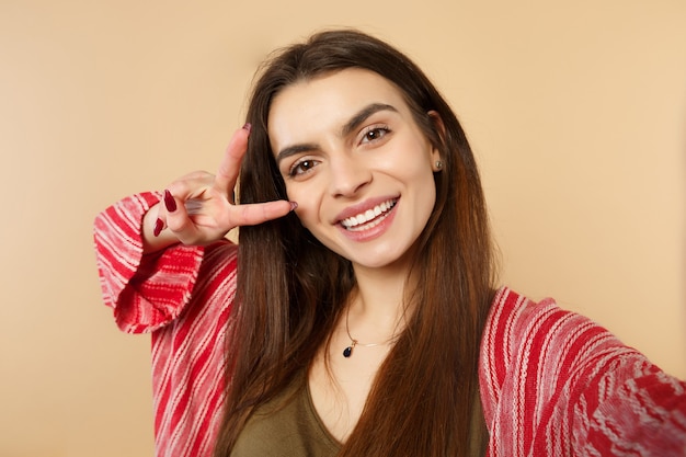 Cierre de tiro selfie de mujer joven alegre en ropa casual mirando a cámara, mostrando el signo de la victoria aislado sobre fondo beige pastel. Personas sinceras emociones, concepto de estilo de vida. Simulacros de espacio de copia.