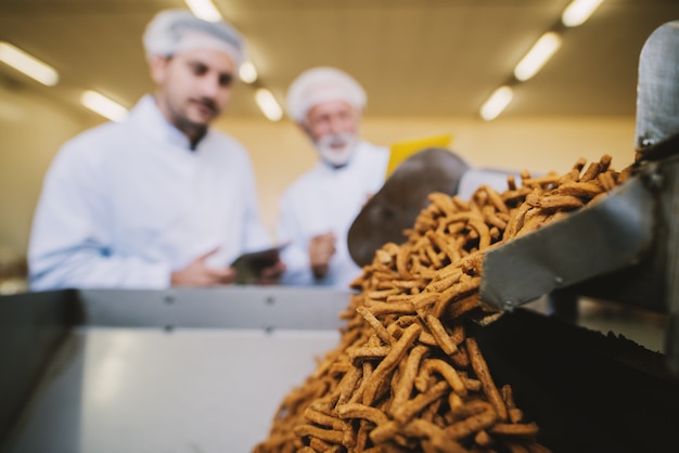 Ciérrenos de bocadillos salados en la línea de producción en la fábrica de alimentos. Imagen borrosa de dos hombres de negocios en ropa esterilizada en segundo plano.