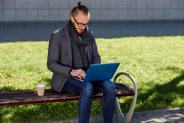 Ciérrese para arriba del hombre joven caucásico con una barba que se sienta en el banco con la computadora portátil y la taza de café. Estudiante sentado en el banco en el parque y usando la computadora portátil.